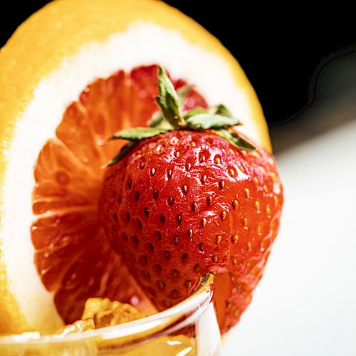 The image shows a close-up of a drink garnish with a slice of orange and a fresh strawberry placed on the rim of a glass.