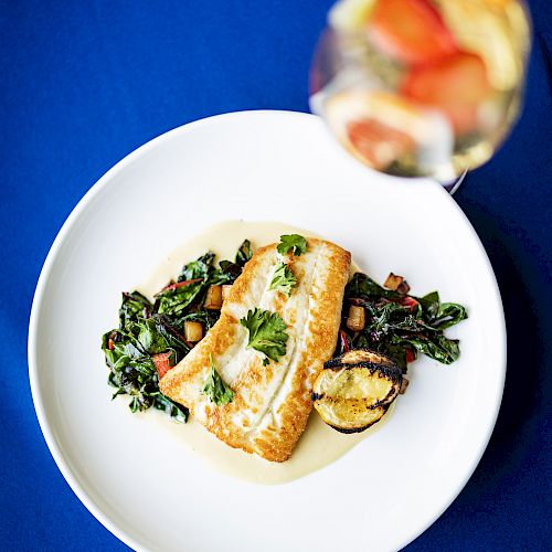 A plate of cooked fish with vegetables and greens is displayed on a blue tablecloth, accompanied by a drink with fruit slices.