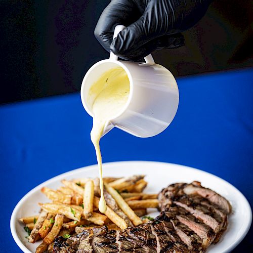 A person wearing a black glove pours sauce from a white jug onto fries and a steak on a white plate, set on a blue tablecloth, ending the sentence.