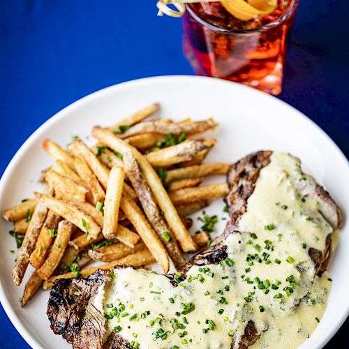 A plate with french fries and steak covered in a creamy sauce, garnished with herbs, beside a cocktail garnished with an orange peel, on a blue tablecloth.