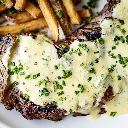 A plate featuring grilled steak topped with a creamy chive sauce, accompanied by a side of seasoned fries on a white plate.