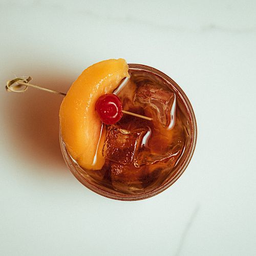 A glass of iced beverage garnished with a peach slice and a cherry, skewered on a toothpick.