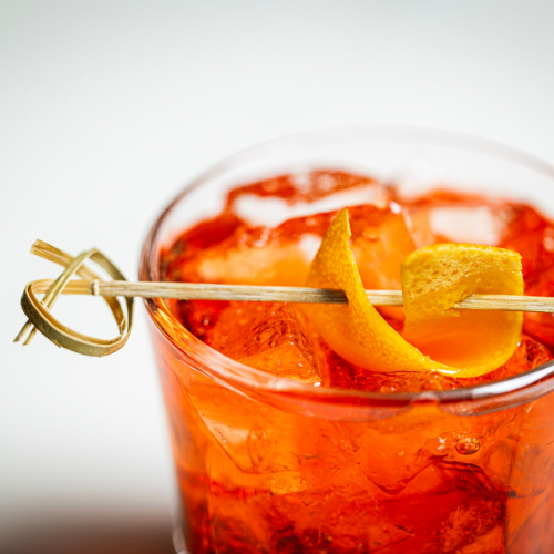 A close-up of an orange cocktail with ice, garnished with a twisted orange peel skewered on a wooden pick.