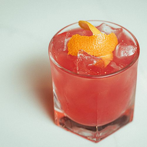 A vibrant red cocktail in a short glass, garnished with an orange peel, served with ice cubes on a light-colored surface.