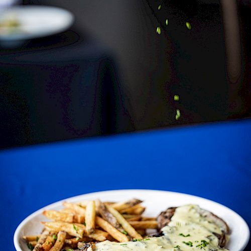 A plate with steak covered in sauce and a side of fries on a blue tablecloth, with a blurry background.