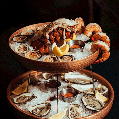 This image shows a two-tiered seafood platter with oysters, shrimp, a crab, lemon wedges, and dipping sauces, garnished on a bed of ice.