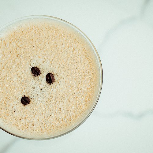 A foamy coffee drink garnished with three coffee beans is pictured from above, placed on a white surface.