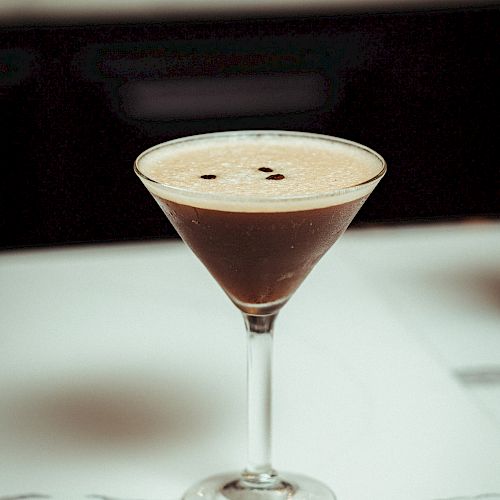 A martini glass with an espresso martini cocktail topped with a couple of coffee beans, placed on a white, marbled surface in a dimly lit setting.