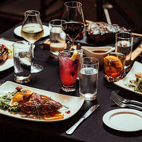 A table set for a meal with various dishes including steak, fish, drinks, and salads on white plates, surrounded by cutlery and glasses.