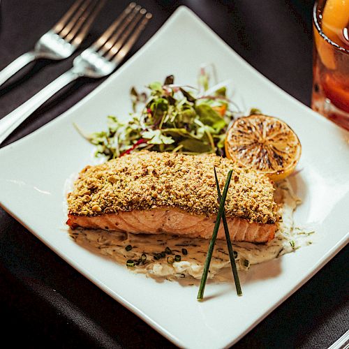 The image shows a plated dish of breaded fish fillet with a lemon slice, greens, and a drink on the side, set on a dining table.