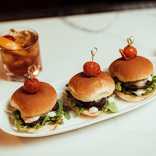 The image shows three mini burgers on a white plate, each topped with a cherry tomato, alongside a drink with an orange slice and a white cloth napkin.