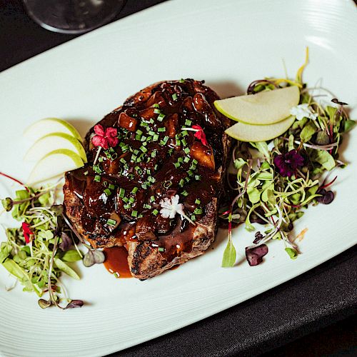 A gourmet dish featuring a glazed meat steak garnished with chopped herbs, microgreens, and apple slices, presented on a white plate.