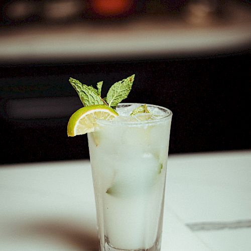 A tall glass of a clear cocktail with ice, garnished with a lemon wedge and sprigs of mint, placed on a marble surface with a blurred background.