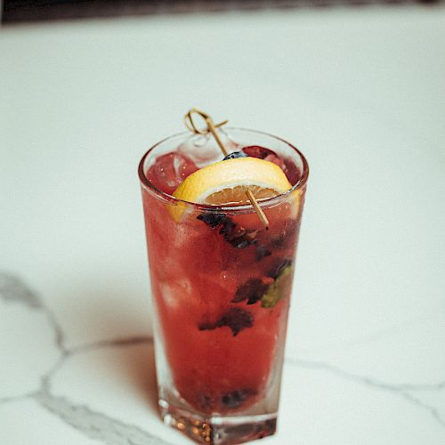 A glass of a red beverage garnished with a lemon slice and fresh herbs, placed on a white marble surface.