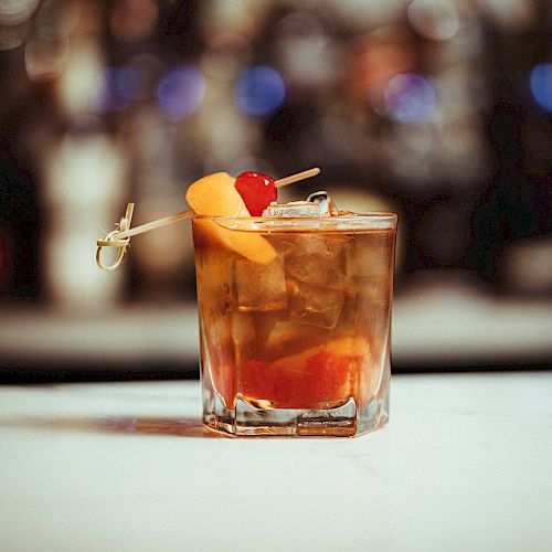 A cocktail garnished with an orange slice and a cherry on a toothpick, served in a glass, set against a blurred background of lights.