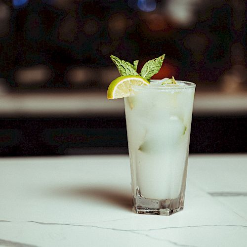 A tall glass of iced drink garnished with a lime wedge and mint leaves on a marble countertop.