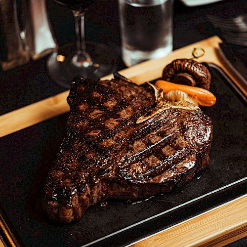 A grilled steak is served on a wooden board with roasted vegetables, accompanied by glasses of water and wine on a dining table.