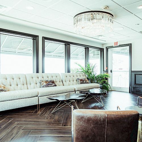 This image shows a modern, bright lounge space with a long couch, coffee tables, plants, and a chandelier, featuring large windows and stylish decor.