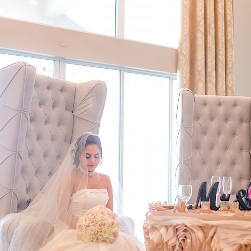 A bride in a white wedding dress and veil sits on a plush chair, holding a flower bouquet, next to a decorated table with a window behind.