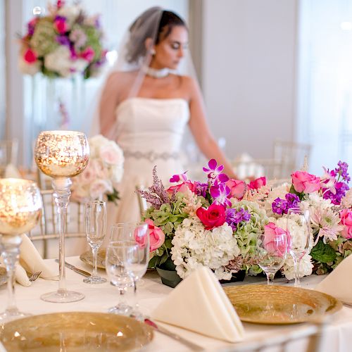 A bride stands behind an elegantly set table with floral centerpieces and gold accents, surrounded by wine glasses and folded napkins.