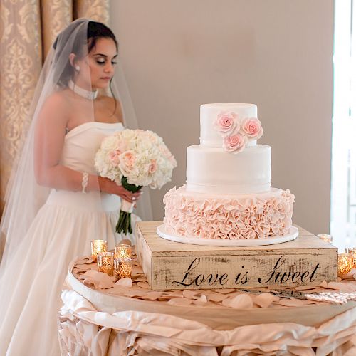 A bride in a white dress and veil holds a bouquet, standing next to a wedding cake on a table with “Love is Sweet” written on it.