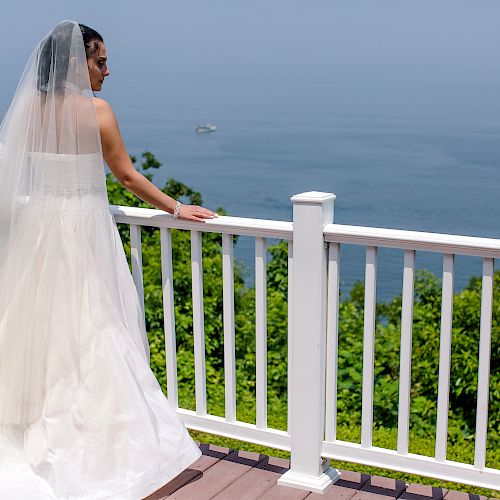 A bride in a wedding dress stands on a balcony, looking out at a scenic ocean view, with greenery and clear skies in the background.