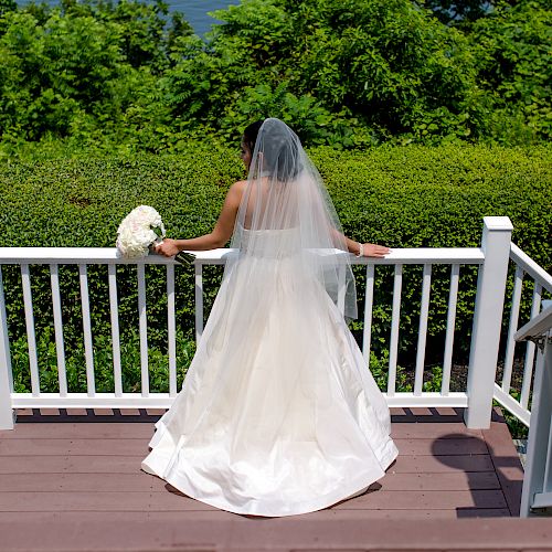 A bride in a white dress with a long veil stands on stairs, holding a bouquet, facing a green garden landscape.