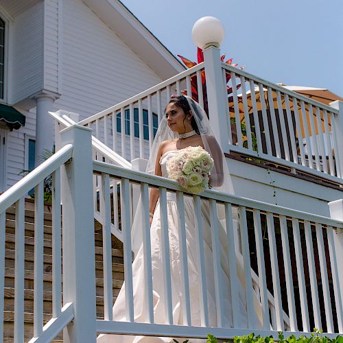 A bride in a white gown holding a bouquet of flowers descends an outdoor staircase with white railings, under a clear sky, near a white building.