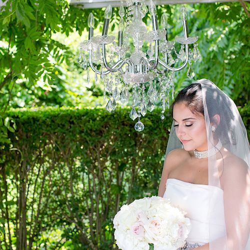 A bride in a white dress stands under a chandelier, holding a bouquet of flowers, surrounded by lush greenery, looking serene.