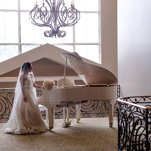 A bride in a white wedding gown and veil stands next to a white grand piano in a spacious, elegant room with large windows and a chandelier.