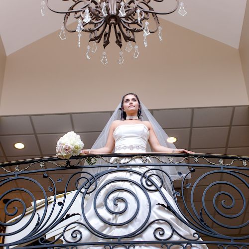 A bride in a white wedding gown stands on a balcony adorned with intricate railings, holding a bouquet, under a chandelier.