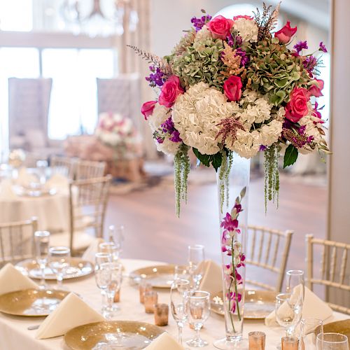 A beautifully set banquet table with a tall, elegant floral arrangement centerpiece featuring pink, white, and purple flowers in a bright, airy room.