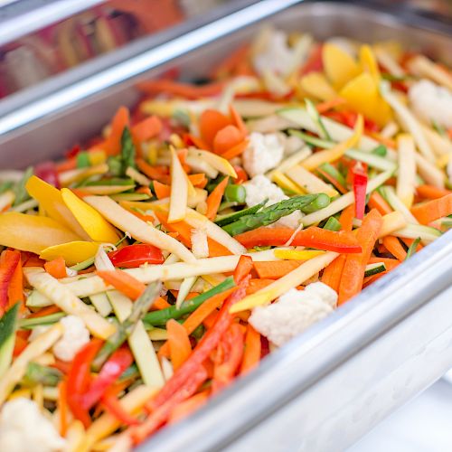 The image shows a stainless steel food tray filled with assorted mixed vegetables such as bell peppers, carrots, asparagus, and cauliflower.