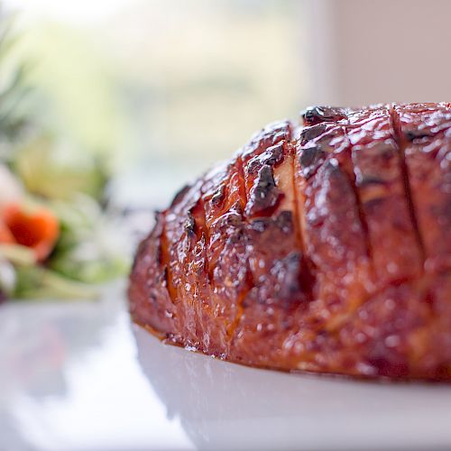 A glazed ham with crisscross cuts is in the foreground on a white plate; blurred vegetables and greens are in the background.